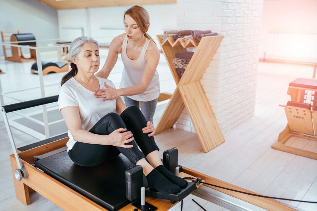 brunette-female-instructor-with-cute-two-braids-hairstyle-consulting-hansdome-beard-man-workout-pilates-on-reformer-practice-in-pilates-studio-working-out-indoor-correcting-beginners-full-length